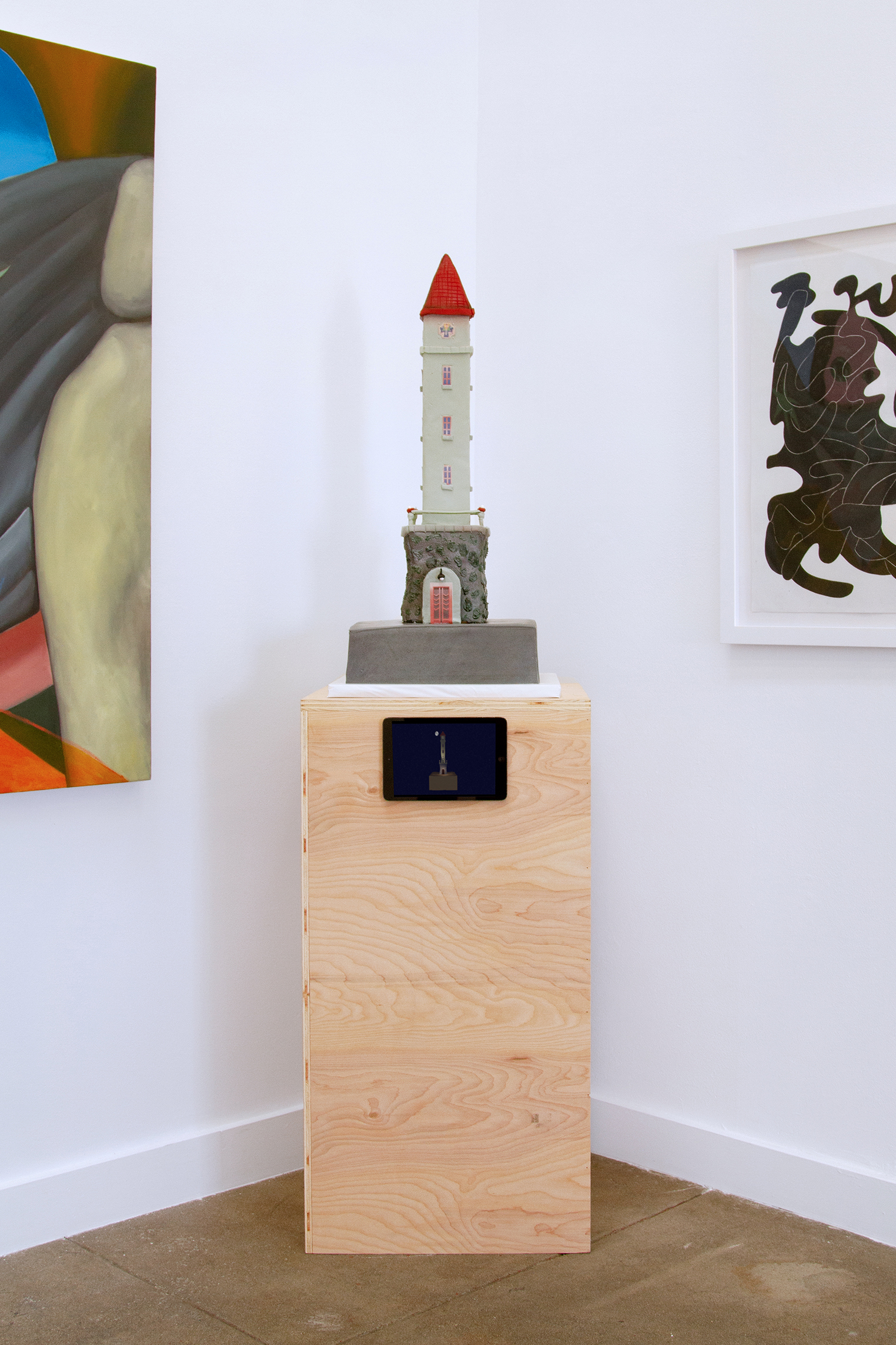 Installation view of a tower shaped cake on a wooden pedestal inbetween two painings